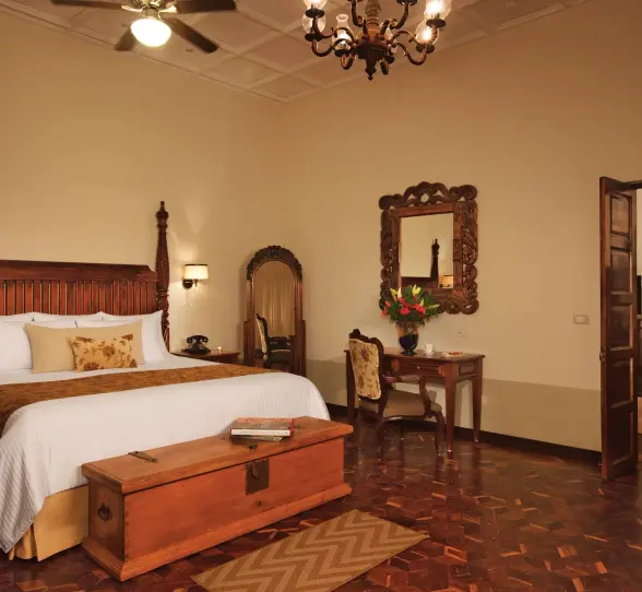 Elegant colonial-style suite with king bed, vintage wood furnishings, and separate sitting area with warm lighting.