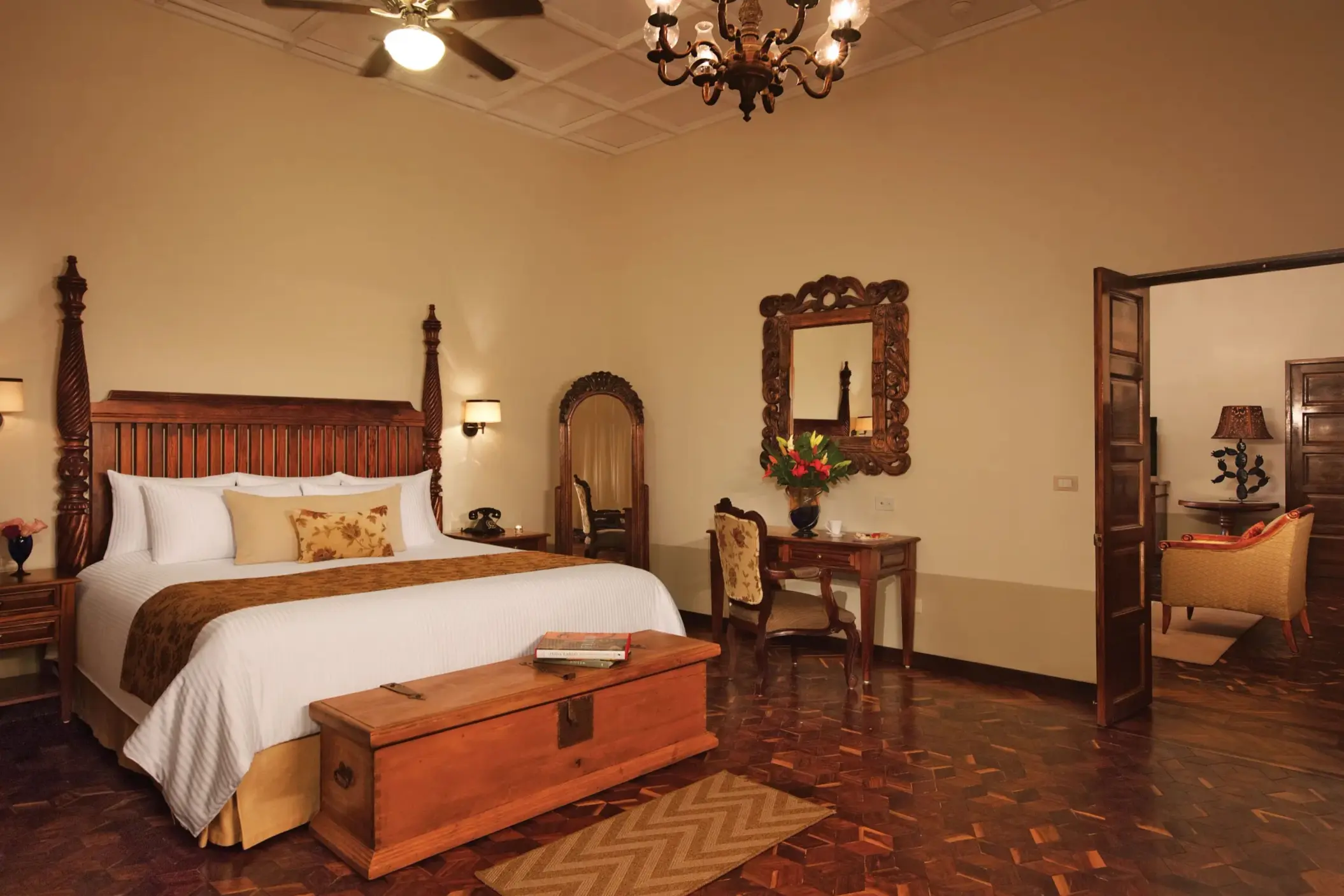 Elegant colonial-style suite with king bed, vintage wood furnishings, and separate sitting area with warm lighting.