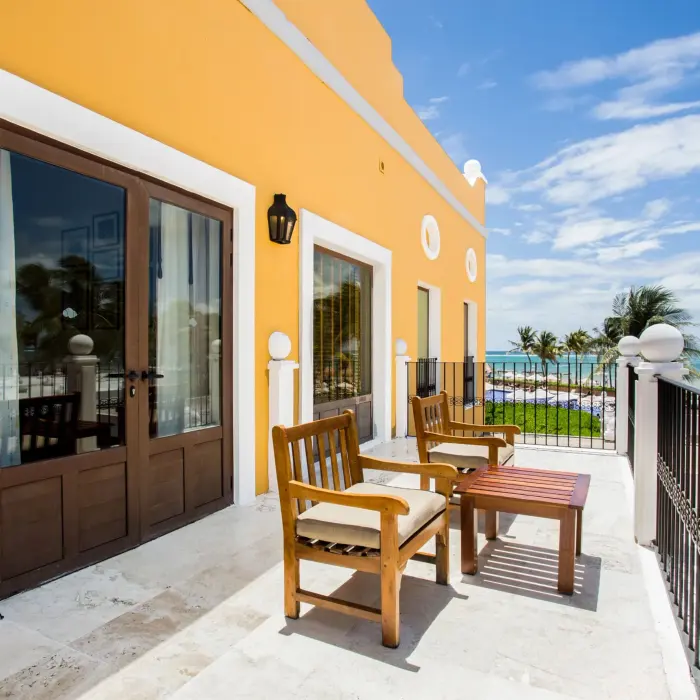 Private balcony with ocean view, wooden chairs, and vibrant yellow exterior walls bathed in Caribbean sunlight.