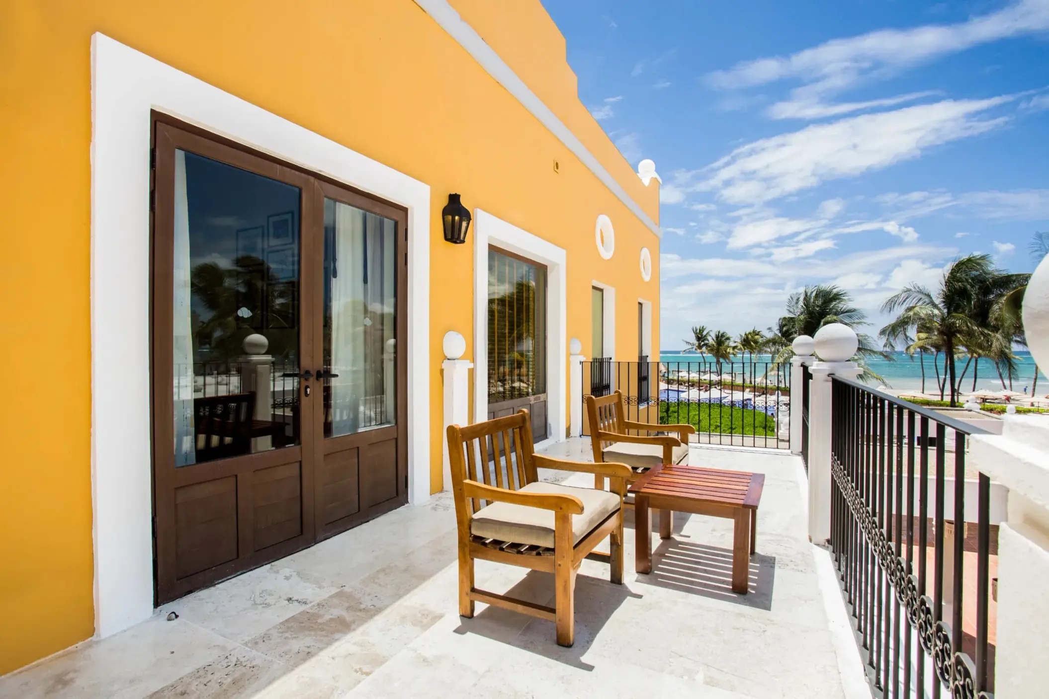 Private balcony with ocean view, wooden chairs, and vibrant yellow exterior walls bathed in Caribbean sunlight.
