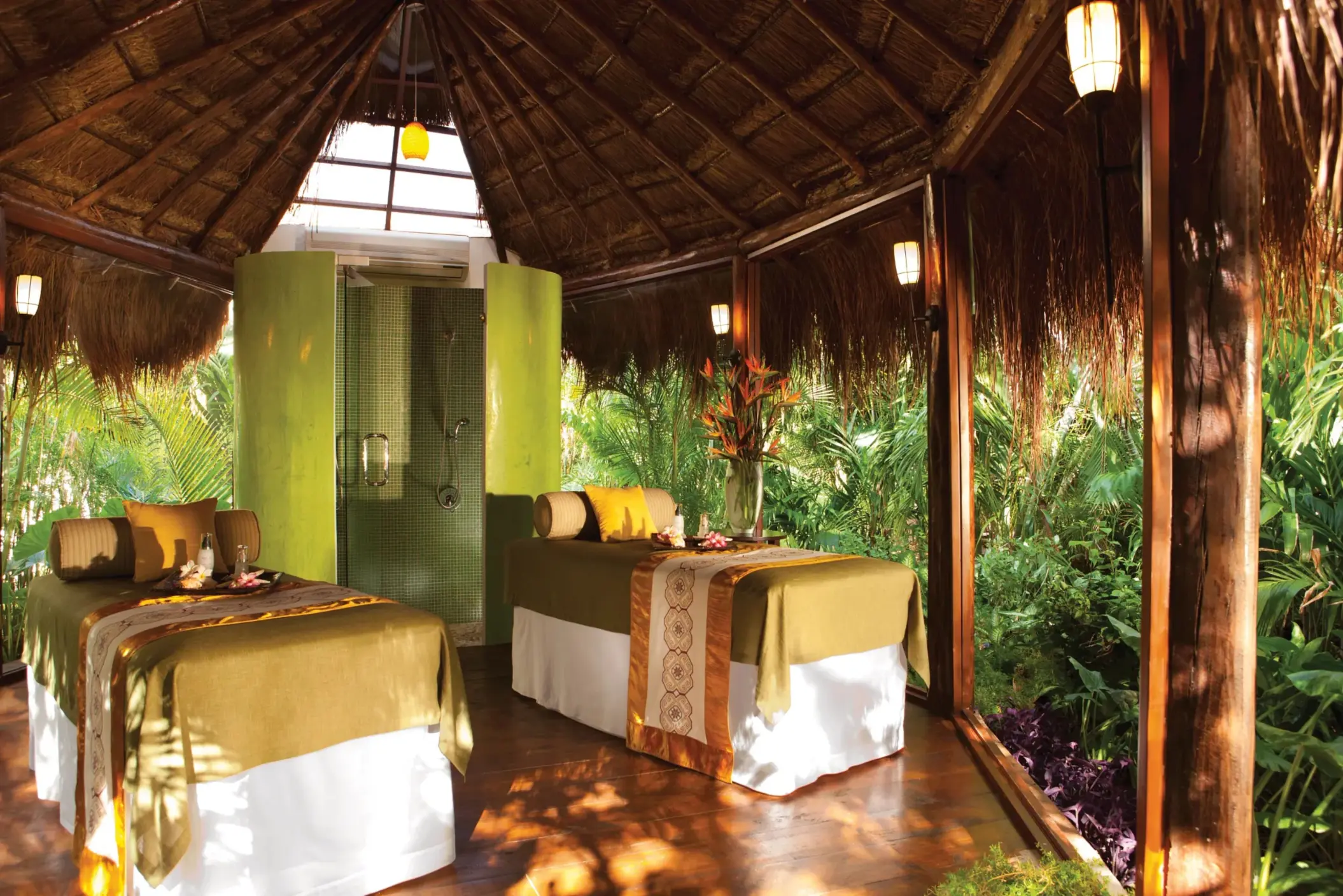 Open-air jungle spa cabin with two massage beds, tropical greenery, and a rainfall shower in the background.