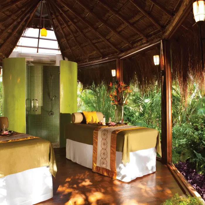Open-air jungle spa cabin with two massage beds, tropical greenery, and a rainfall shower in the background.