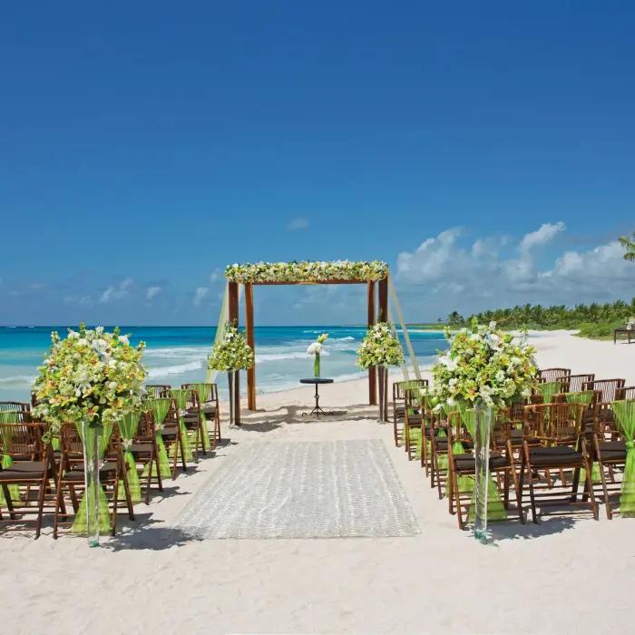 Daytime beach wedding ceremony with wooden chairs, flower arrangements, and oceanfront altar under blue skies.