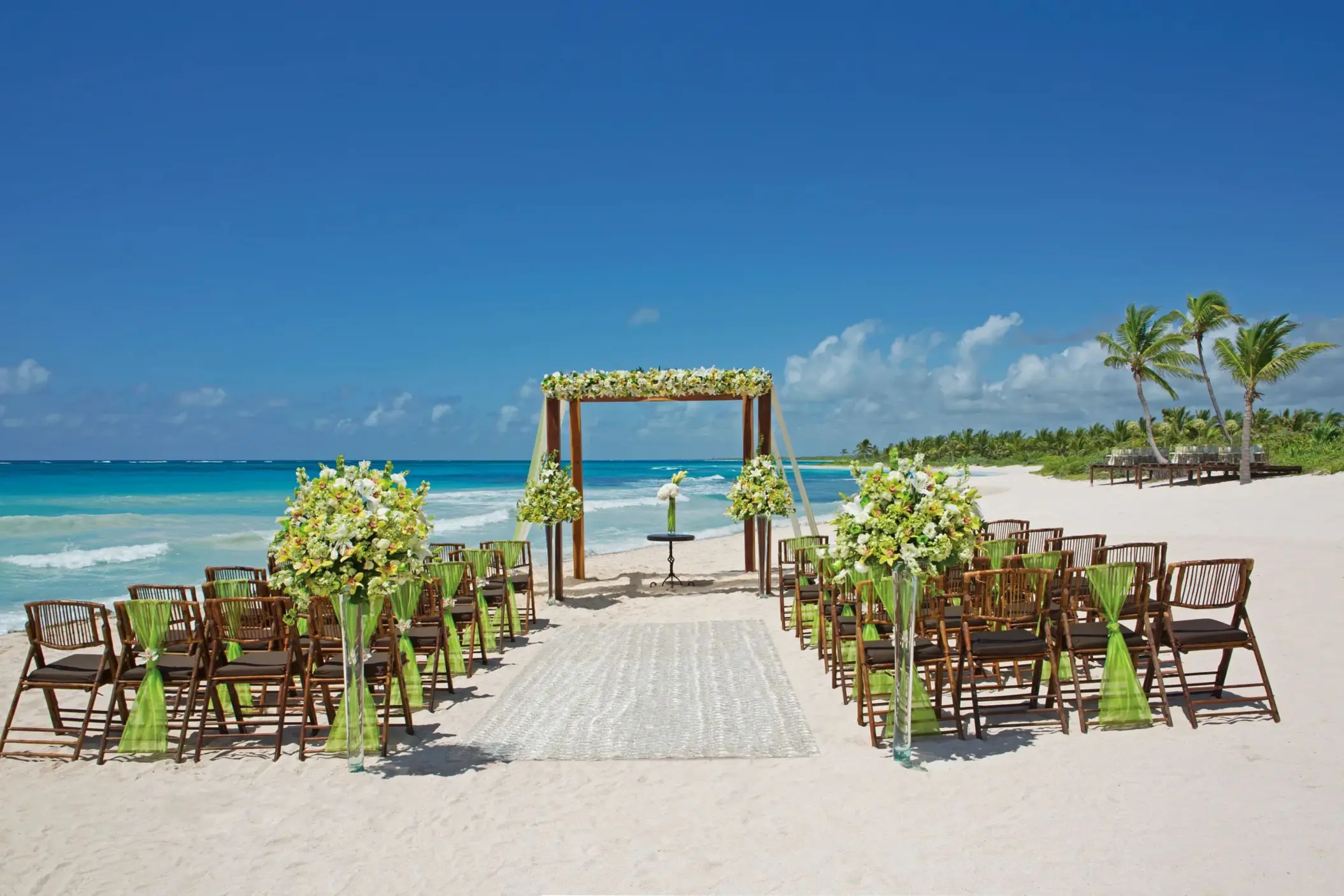 Daytime beach wedding ceremony with wooden chairs, flower arrangements, and oceanfront altar under blue skies.