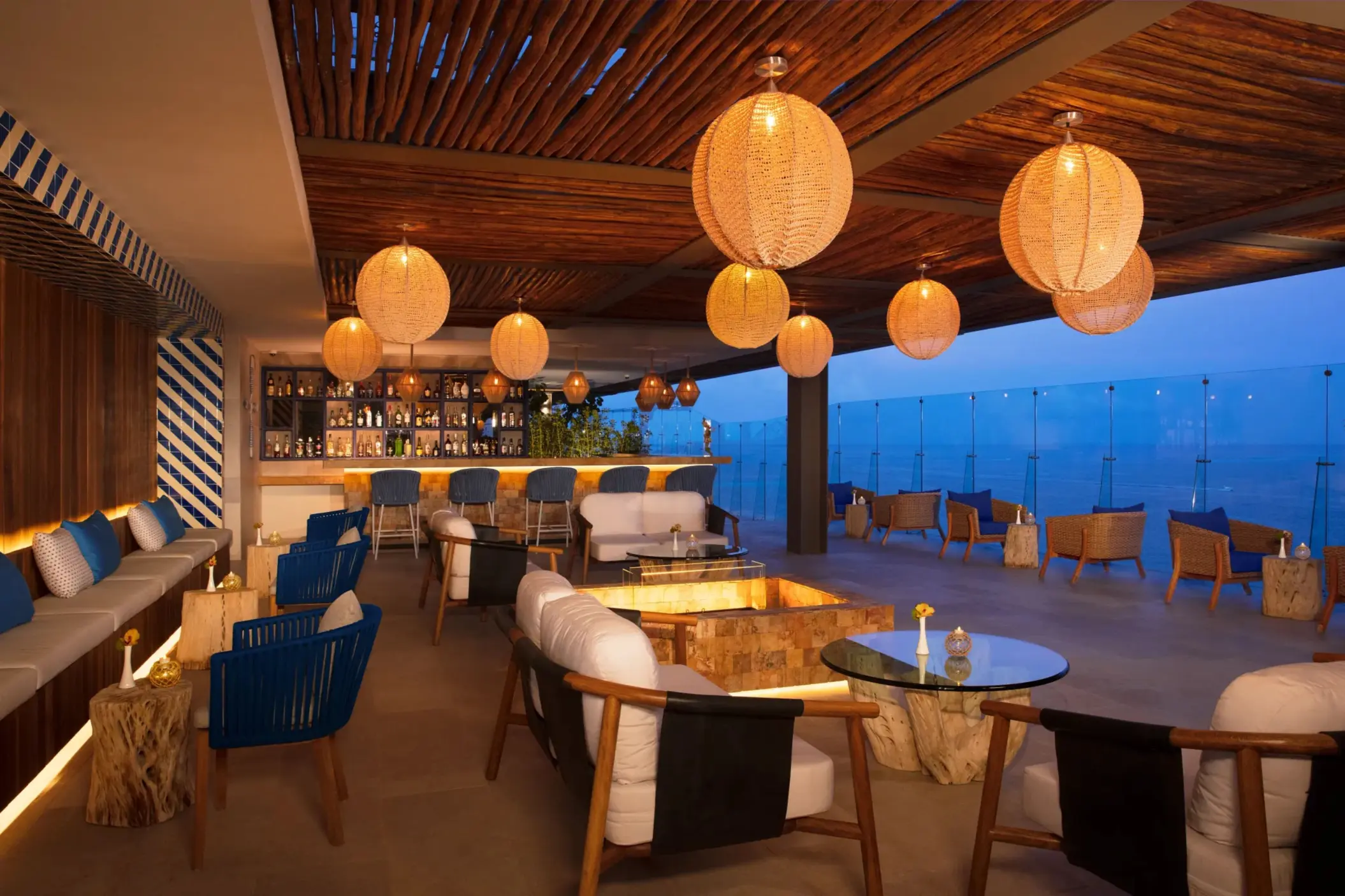 Elegant rooftop bar with cozy lounge seating, hanging lights, and sweeping views of the ocean.