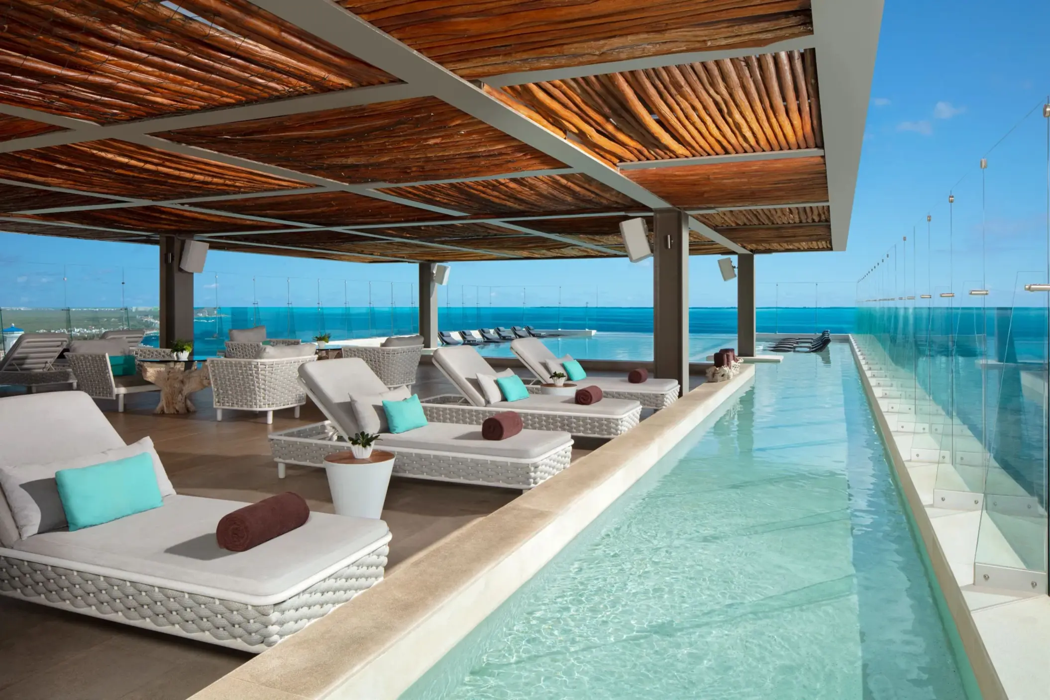 Rooftop poolside lounge with woven sunbeds, wood ceiling, and glass panels overlooking the sea.