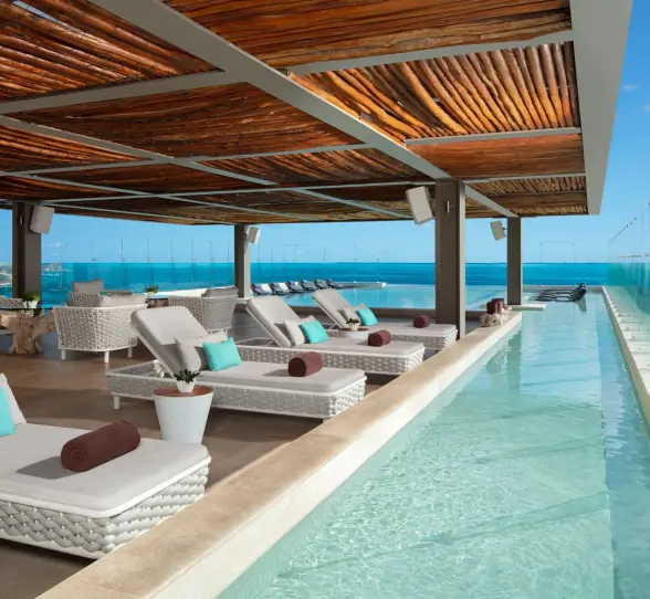 Rooftop poolside lounge with woven sunbeds, wood ceiling, and glass panels overlooking the sea.