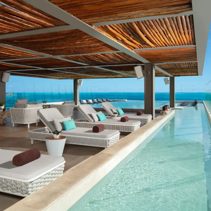 Rooftop poolside lounge with woven sunbeds, wood ceiling, and glass panels overlooking the sea.