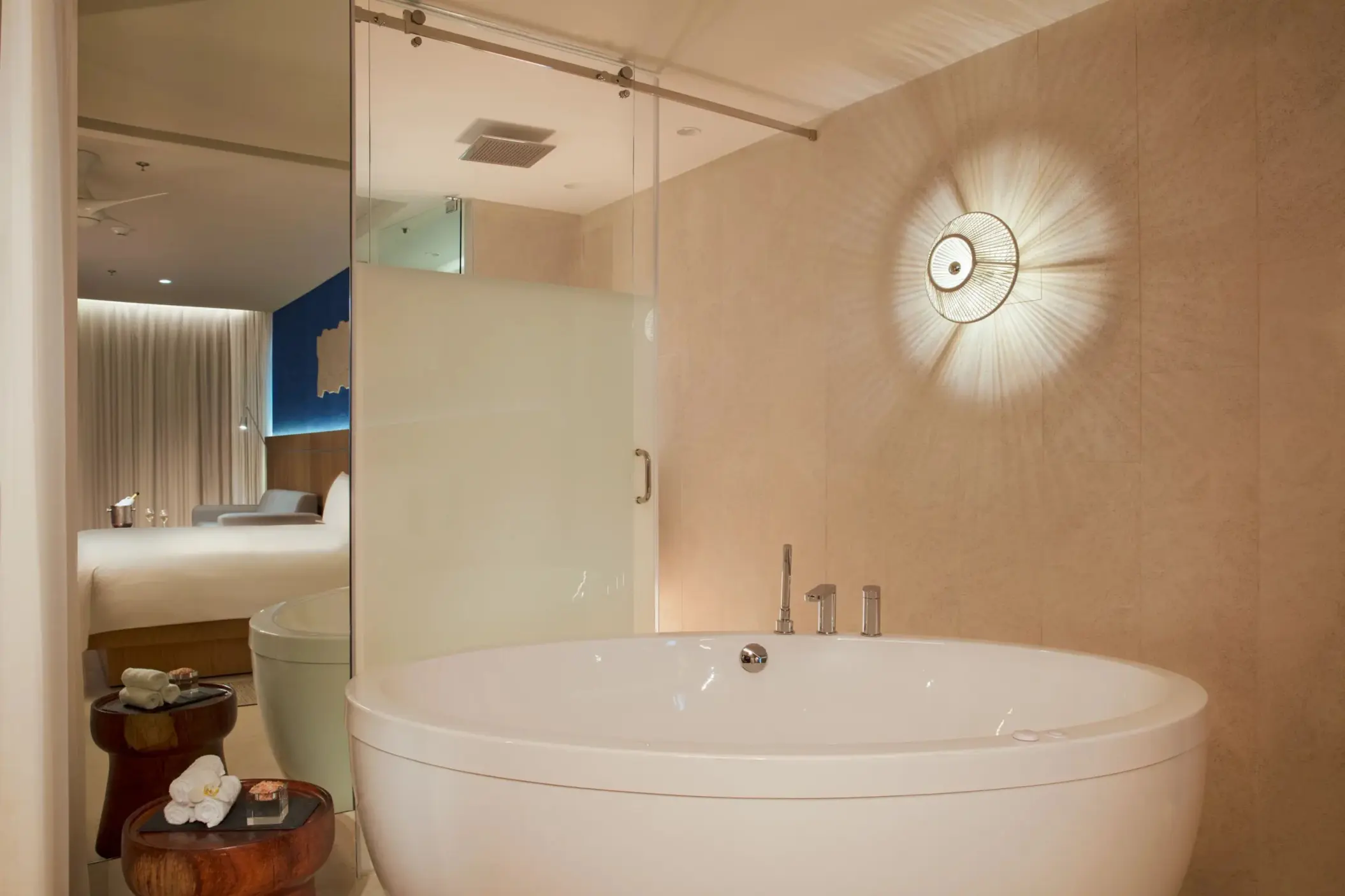 Large round soaking tub with modern wall light and peek into suite’s serene bedroom through glass partition.