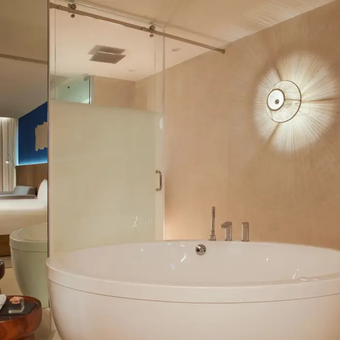 Large round soaking tub with modern wall light and peek into suite’s serene bedroom through glass partition.