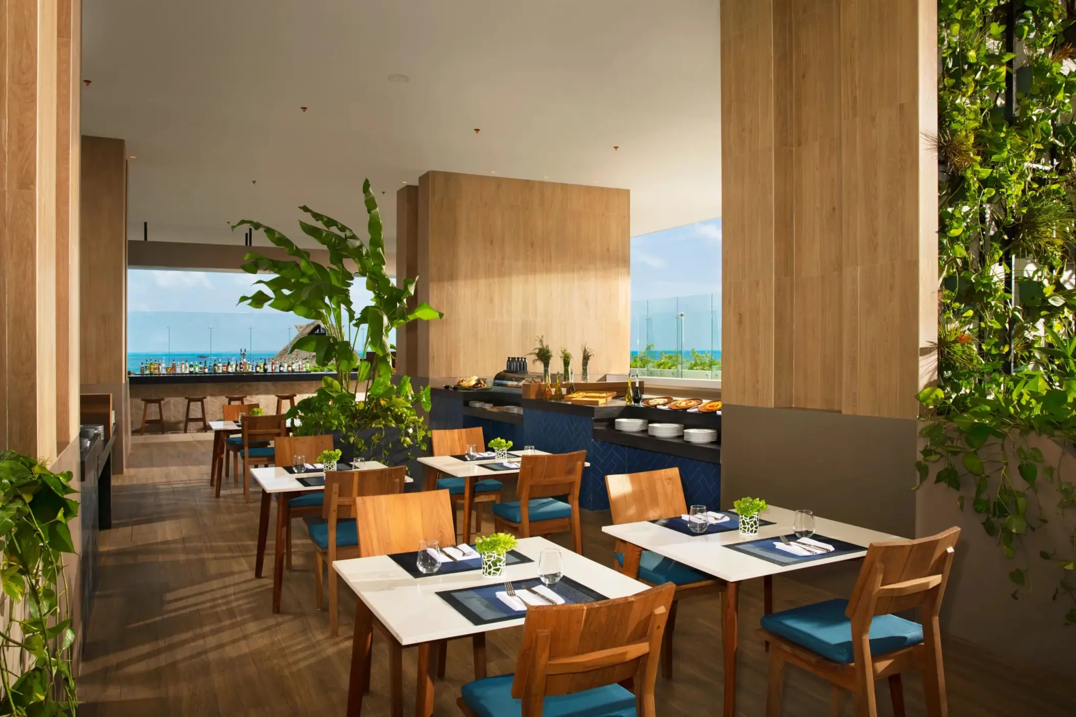 Open-air La Plaza dining area with buffet setup, natural wood decor, and green plants.