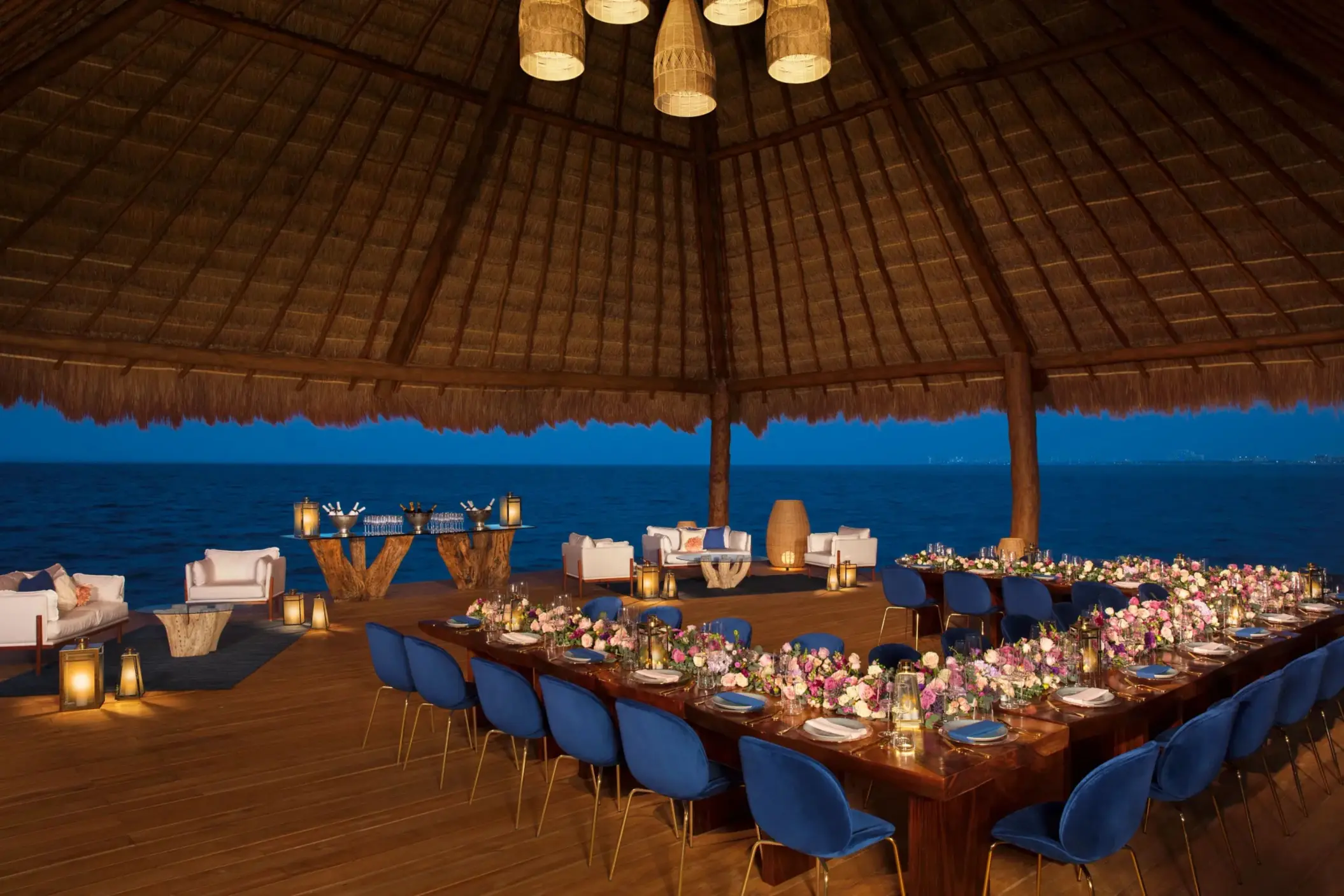 Evening wedding reception setup under a palapa with ocean views and romantic lighting.