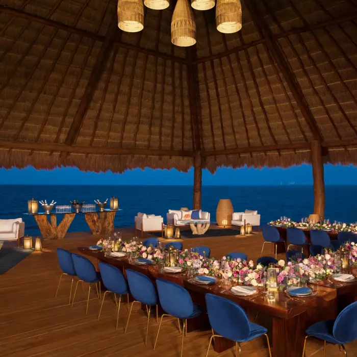 Evening wedding reception setup under a palapa with ocean views and romantic lighting.