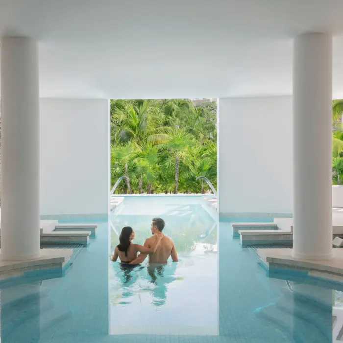 Couple enjoying a hydrotherapy pool session with views of lush jungle through wide open spa windows.