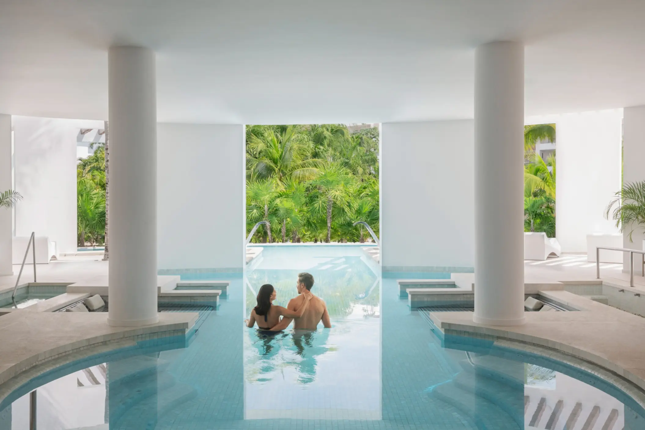 Couple enjoying a hydrotherapy pool session with views of lush jungle through wide open spa windows.