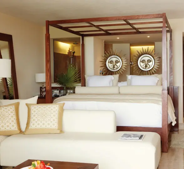 Bright, cozy suite with wooden canopy bed, sun-themed wall art, cream seating, and soft neutral tones