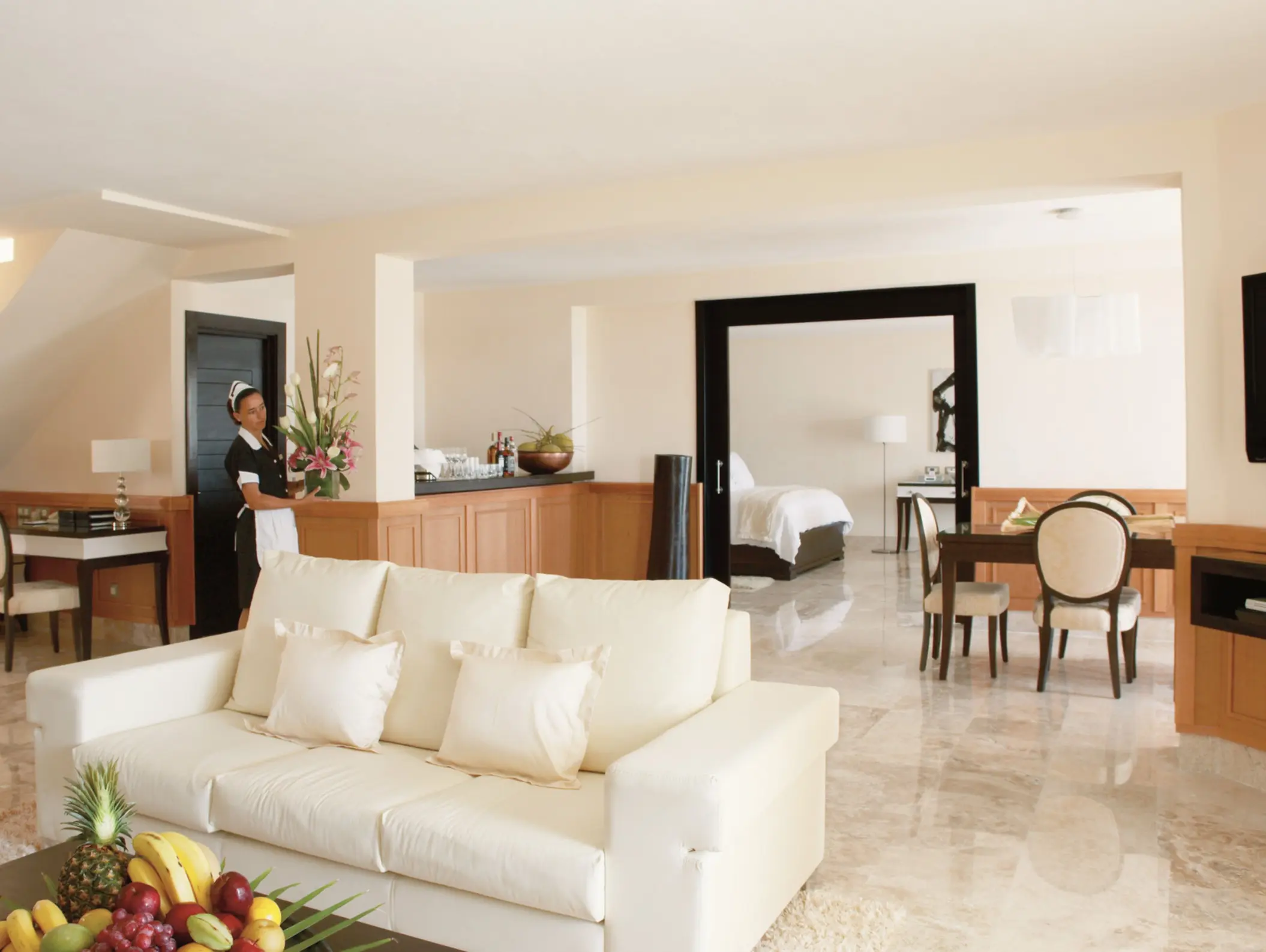 Spacious luxury suite with open layout, cream couch, dining area, fresh flowers, and a stocked minibar.