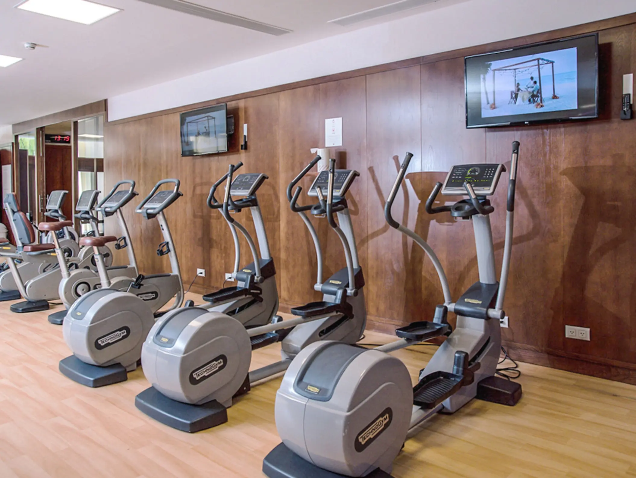 Modern resort gym with Technogym cardio machines, wood paneling, and TVs playing beachside workout videos.