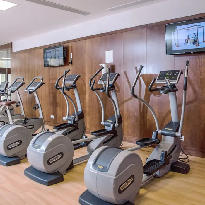 Modern resort gym with Technogym cardio machines, wood paneling, and TVs playing beachside workout videos.