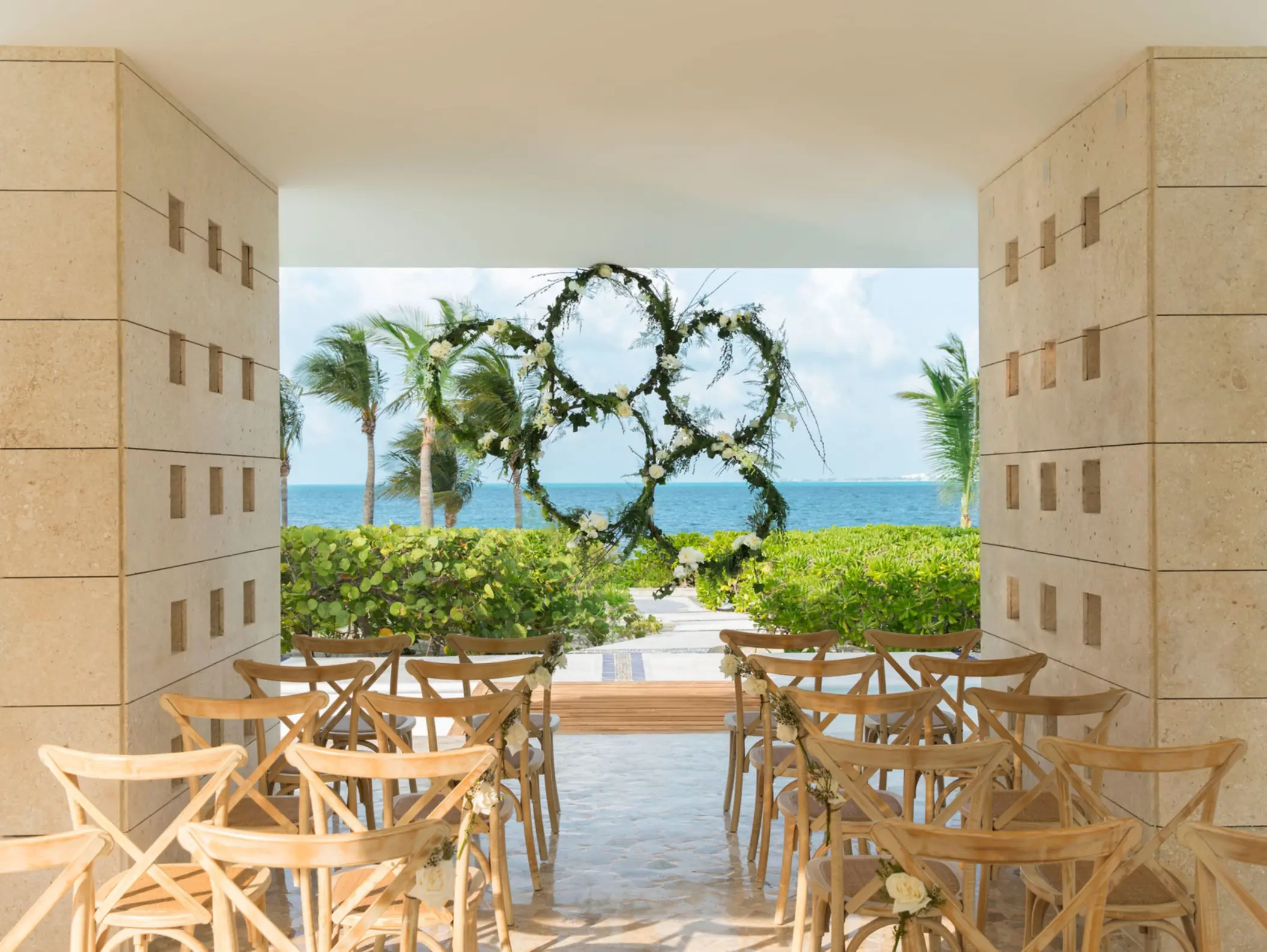 Romantic beachfront wedding gazebo framed by greenery, wooden chairs, and a floral circular arch facing the ocean.