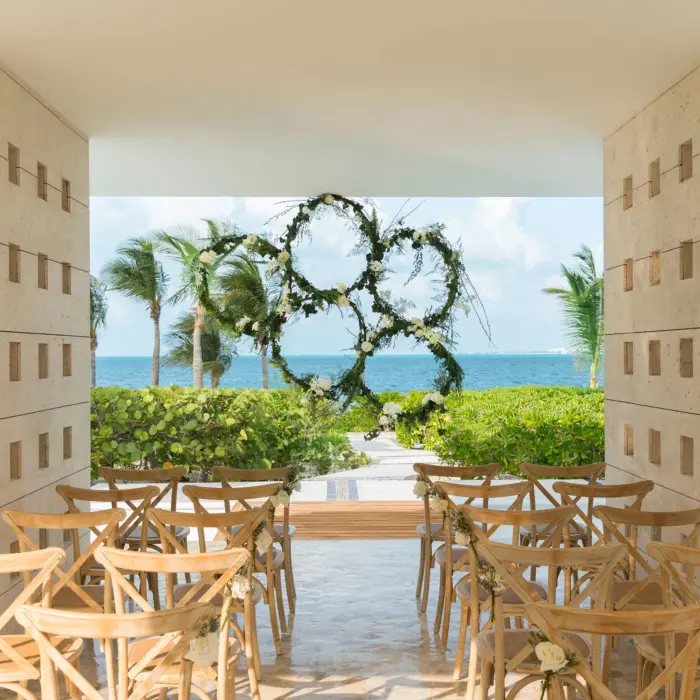 Romantic beachfront wedding gazebo framed by greenery, wooden chairs, and a floral circular arch facing the ocean.