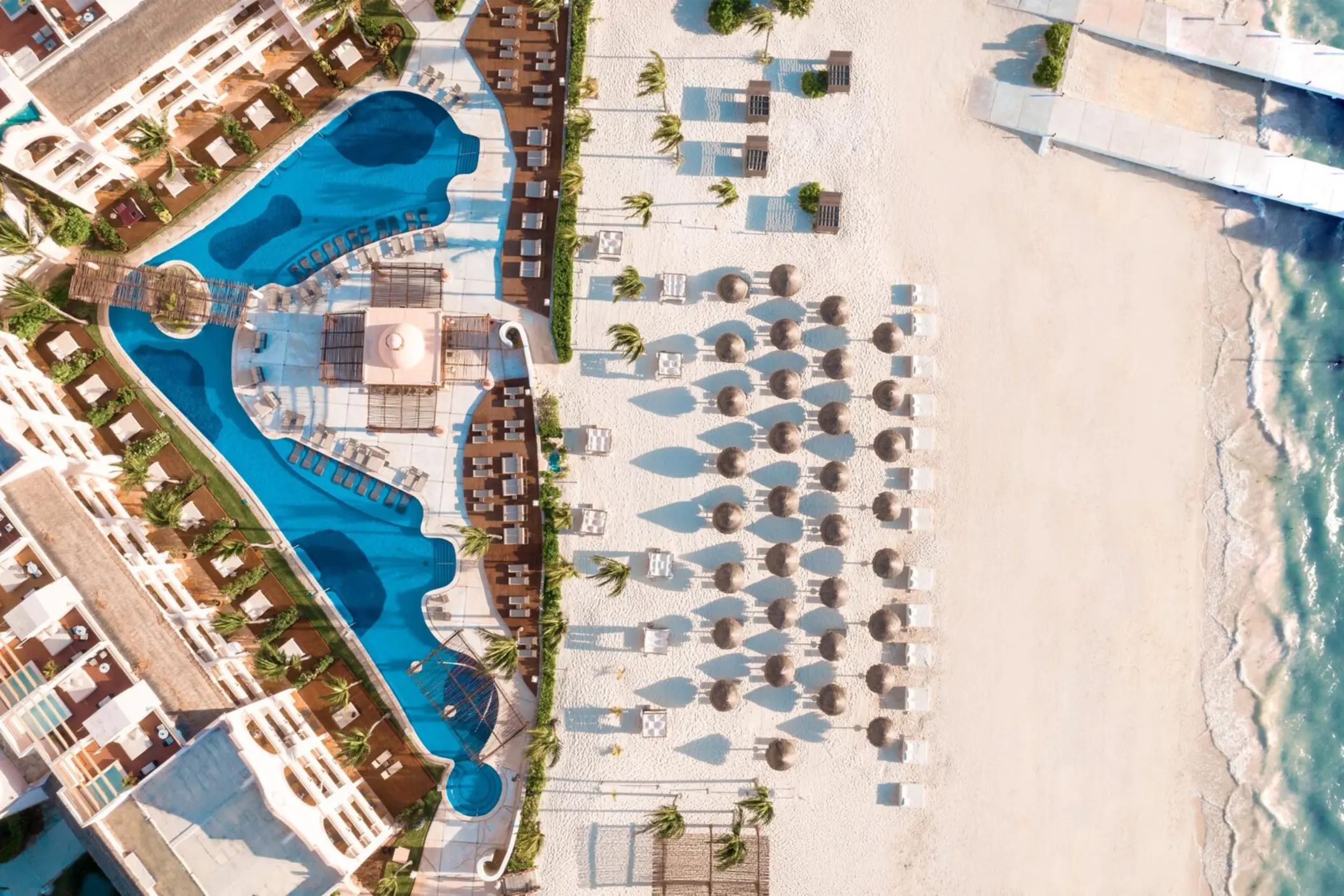 Aerial view of beachfront resort with pools, palapas, lounge chairs, and white sand meeting the turquoise Caribbean.