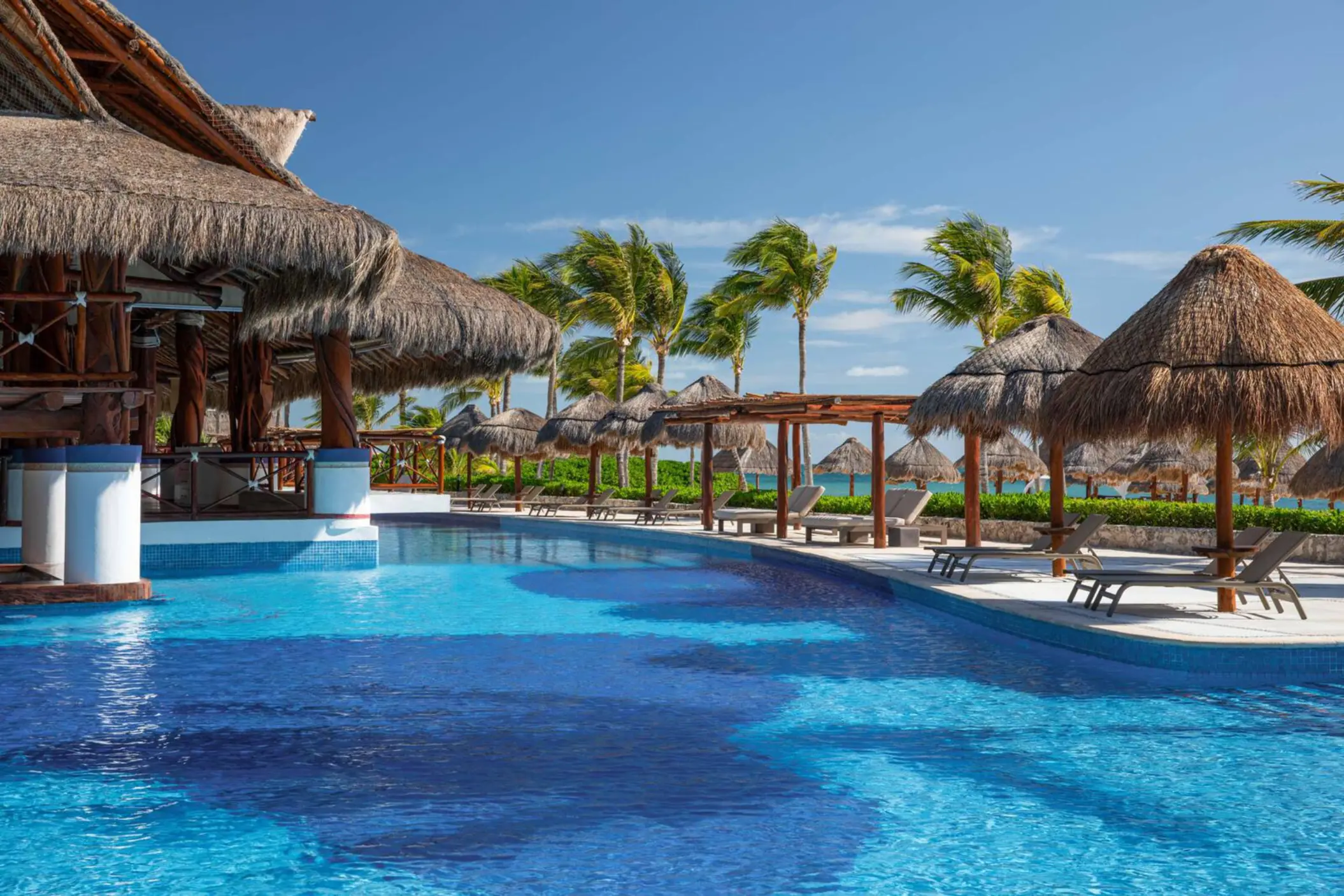 Tranquil resort pool with swim-up bar, palapa-covered loungers, and palm trees swaying under a clear blue sky.