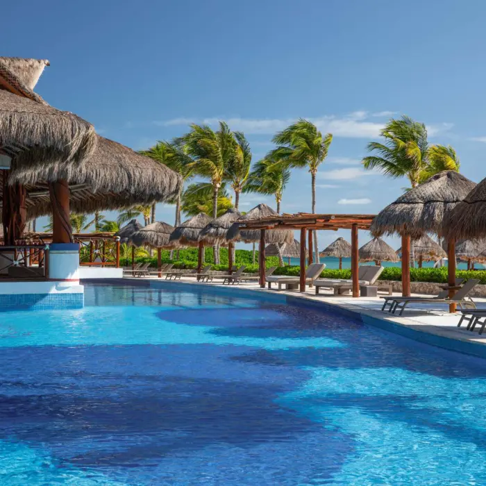 Tranquil resort pool with swim-up bar, palapa-covered loungers, and palm trees swaying under a clear blue sky.
