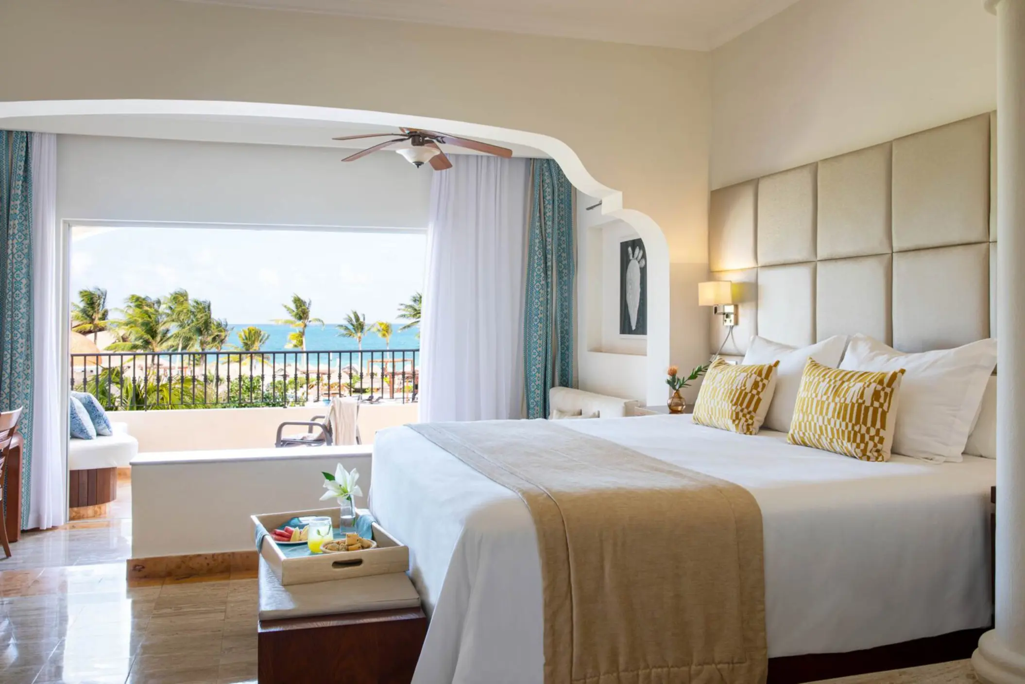 Bright ocean view suite with king bed, cozy lounge, neutral decor, and balcony overlooking palm trees and sea.