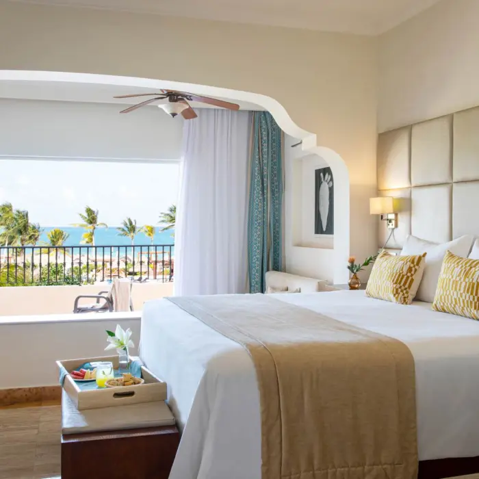Bright ocean view suite with king bed, cozy lounge, neutral decor, and balcony overlooking palm trees and sea.