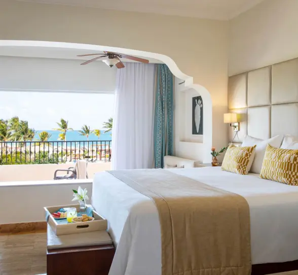 Bright ocean view suite with king bed, cozy lounge, neutral decor, and balcony overlooking palm trees and sea.