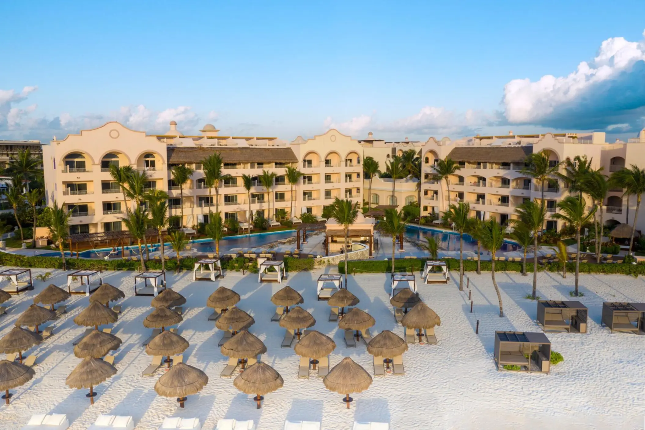 Aerial view of resort facing the beach, lined with lounge chairs and palapas on white sand with palm trees throughout.
