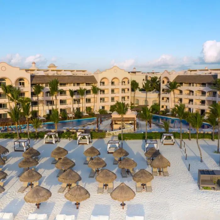 Aerial view of resort facing the beach, lined with lounge chairs and palapas on white sand with palm trees throughout.