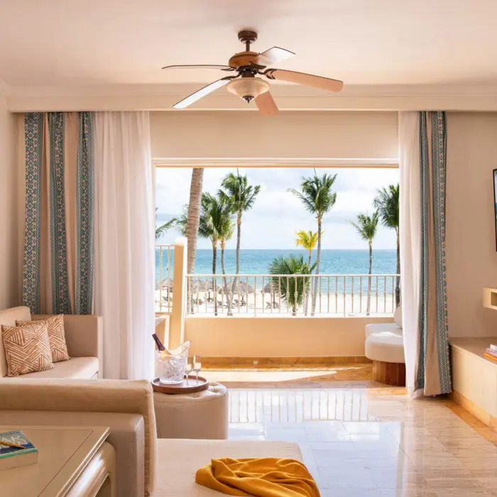 Romantic honeymoon suite with beach views, plush seating, champagne, and tropical scenery framed by open curtains.