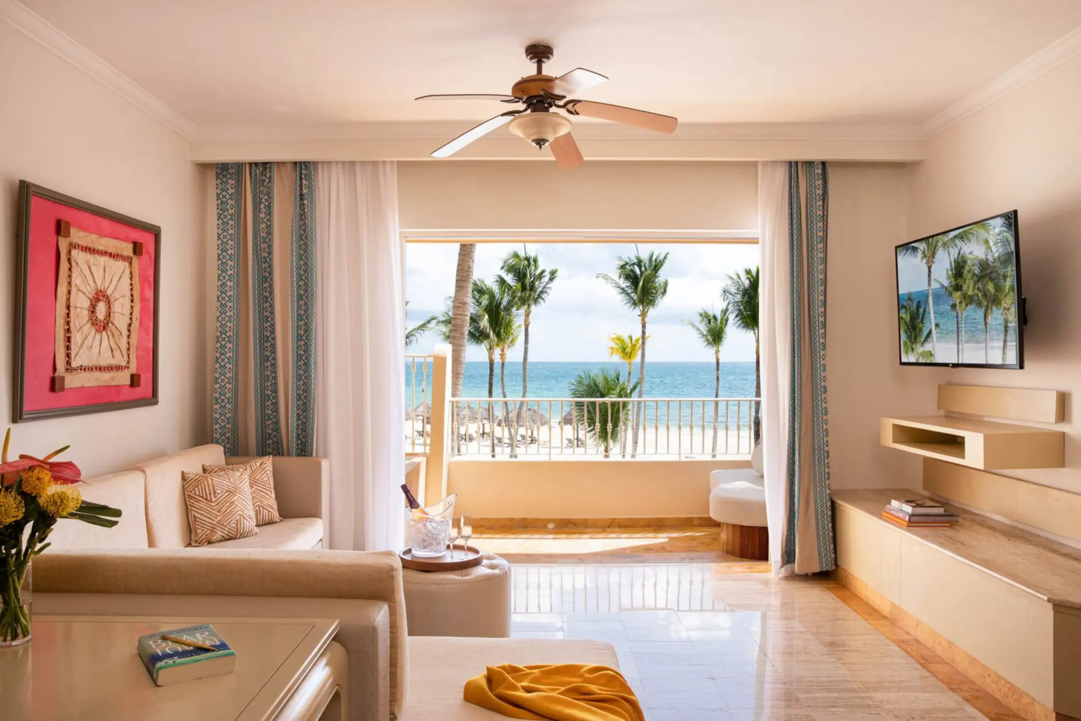 Romantic honeymoon suite with beach views, plush seating, champagne, and tropical scenery framed by open curtains.