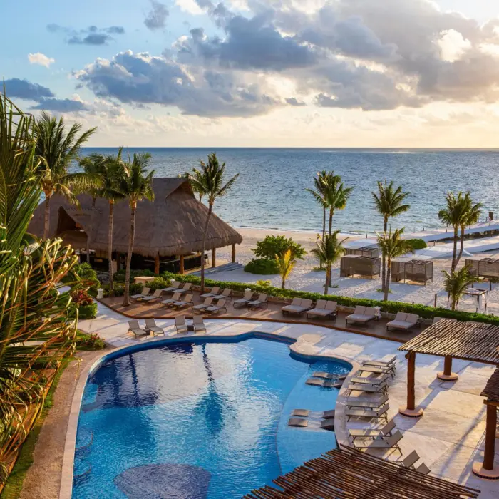 Scenic poolside view of beachfront resort with loungers, palm trees, and sunset over the ocean horizon.