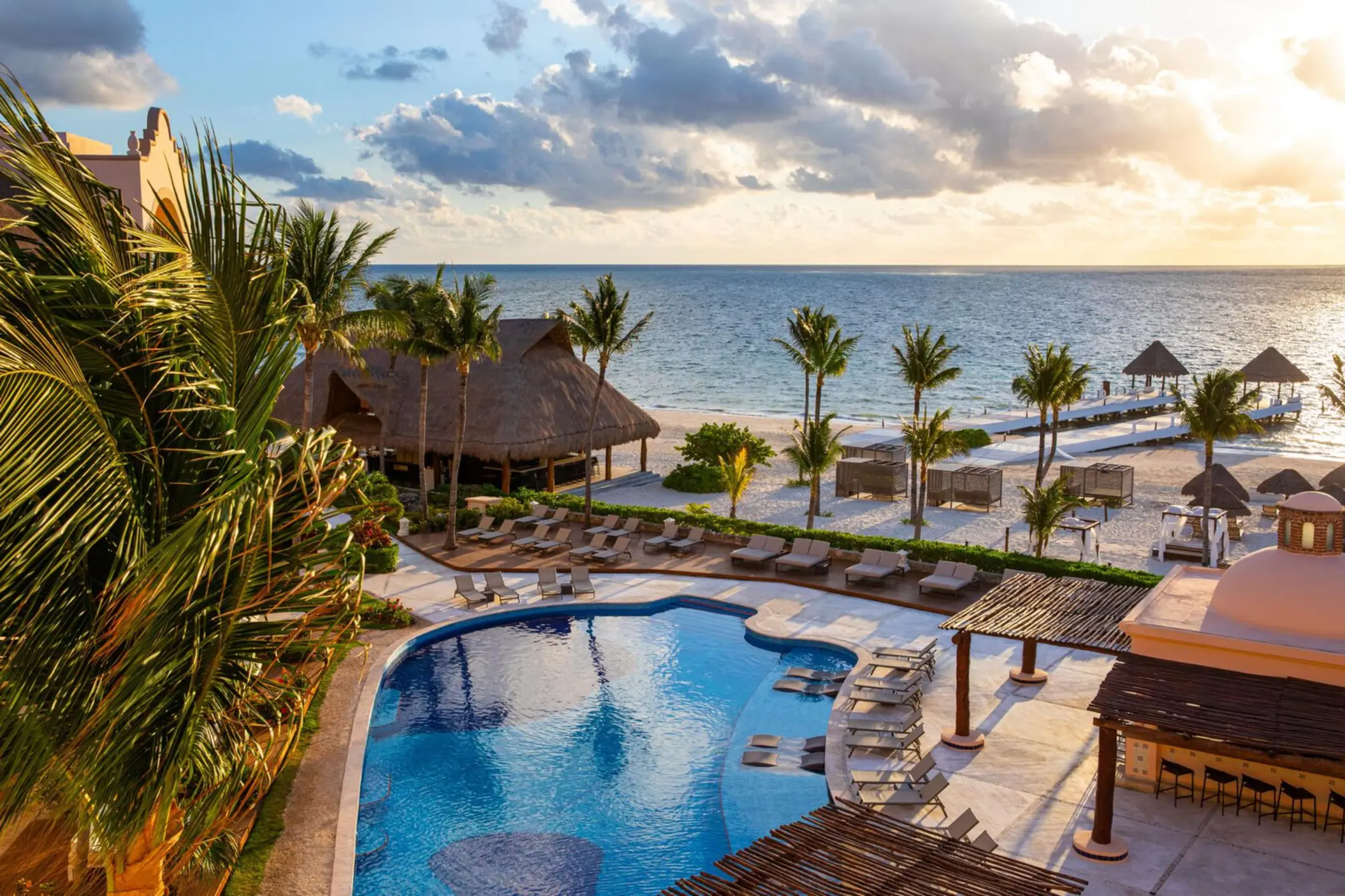 Scenic poolside view of beachfront resort with loungers, palm trees, and sunset over the ocean horizon.