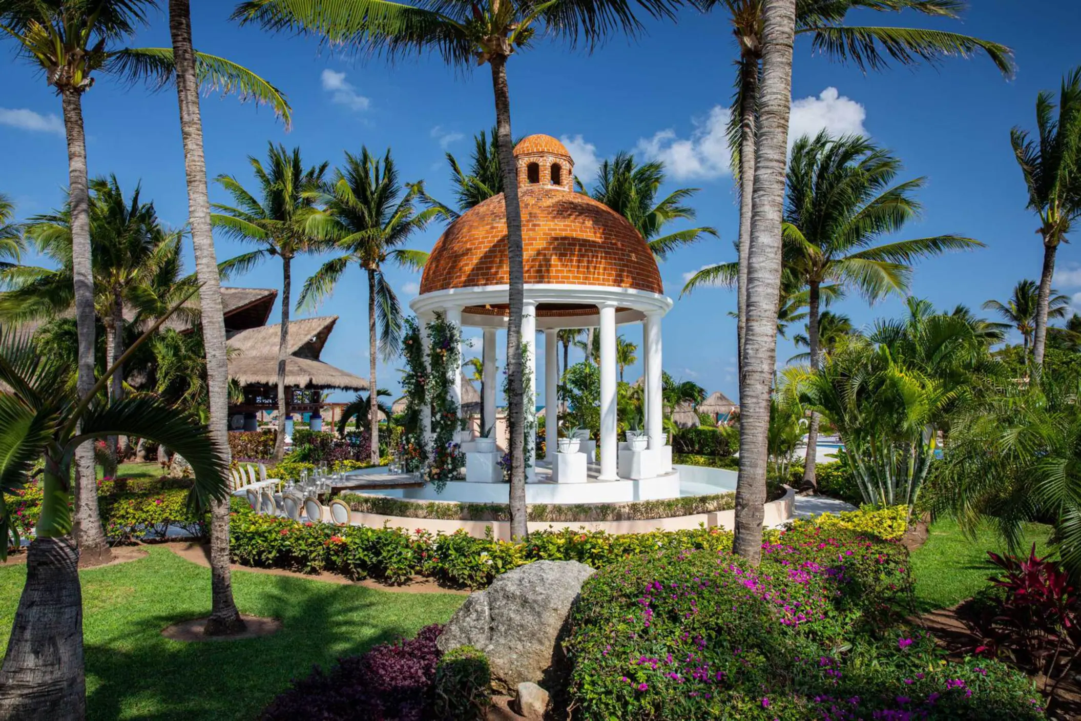 Romantic white wedding gazebo with domed roof surrounded by tropical palms and vibrant garden flowers.