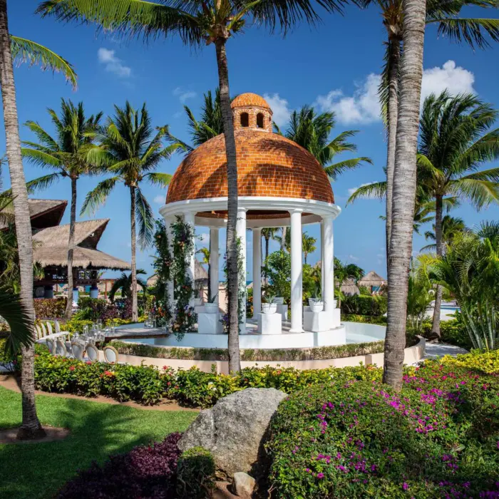 Romantic white wedding gazebo with domed roof surrounded by tropical palms and vibrant garden flowers.