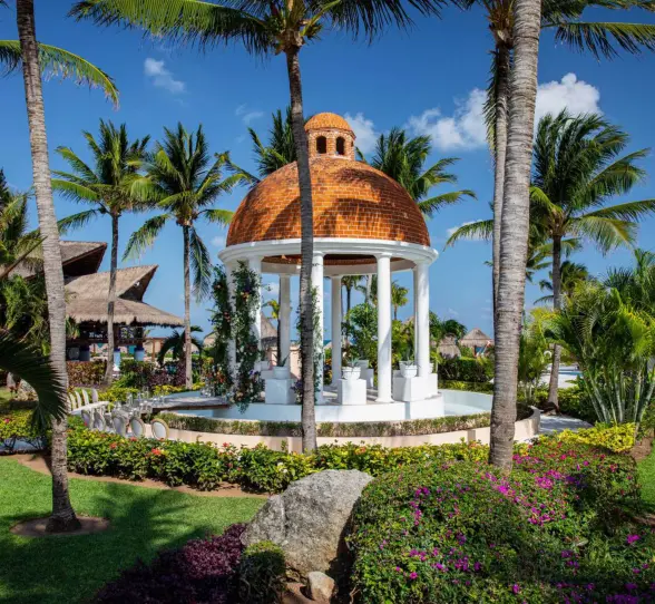 Romantic white wedding gazebo with domed roof surrounded by tropical palms and vibrant garden flowers.