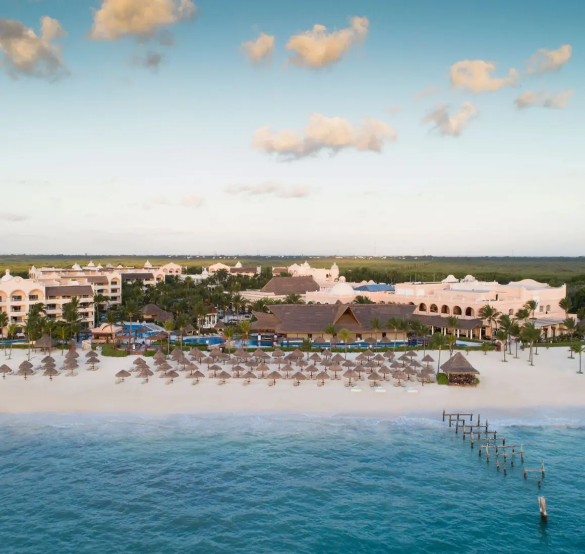 Aerial view of beachfront resort with palapa umbrellas, lush palms, and calm turquoise waters at sunset.