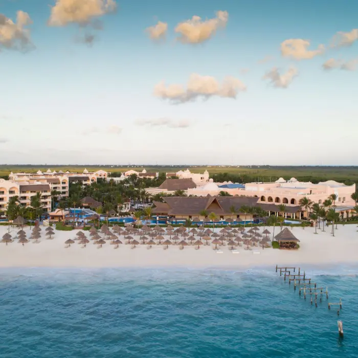 Aerial view of beachfront resort with palapa umbrellas, lush palms, and calm turquoise waters at sunset.