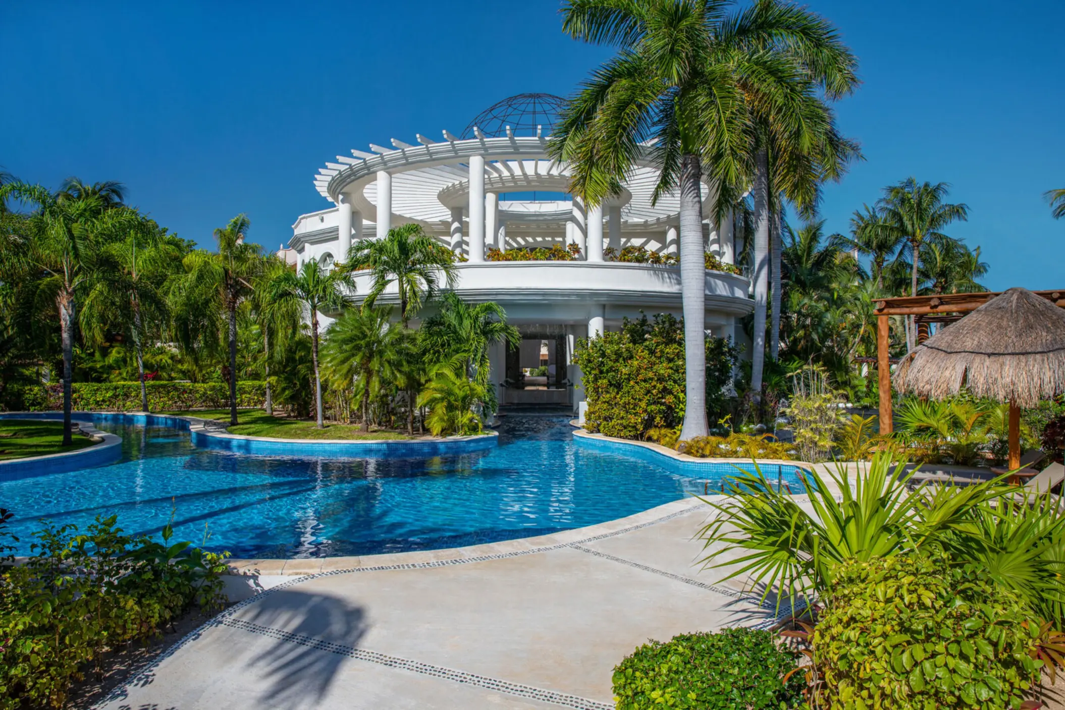 Elegant white-columned pavilion surrounded by tropical gardens and a tranquil pool under a bright blue sky.
