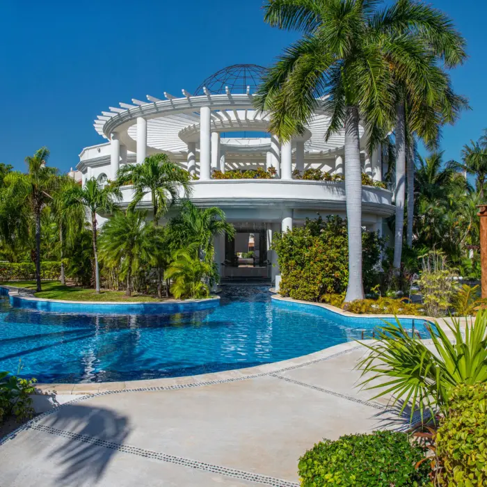 Elegant white-columned pavilion surrounded by tropical gardens and a tranquil pool under a bright blue sky.