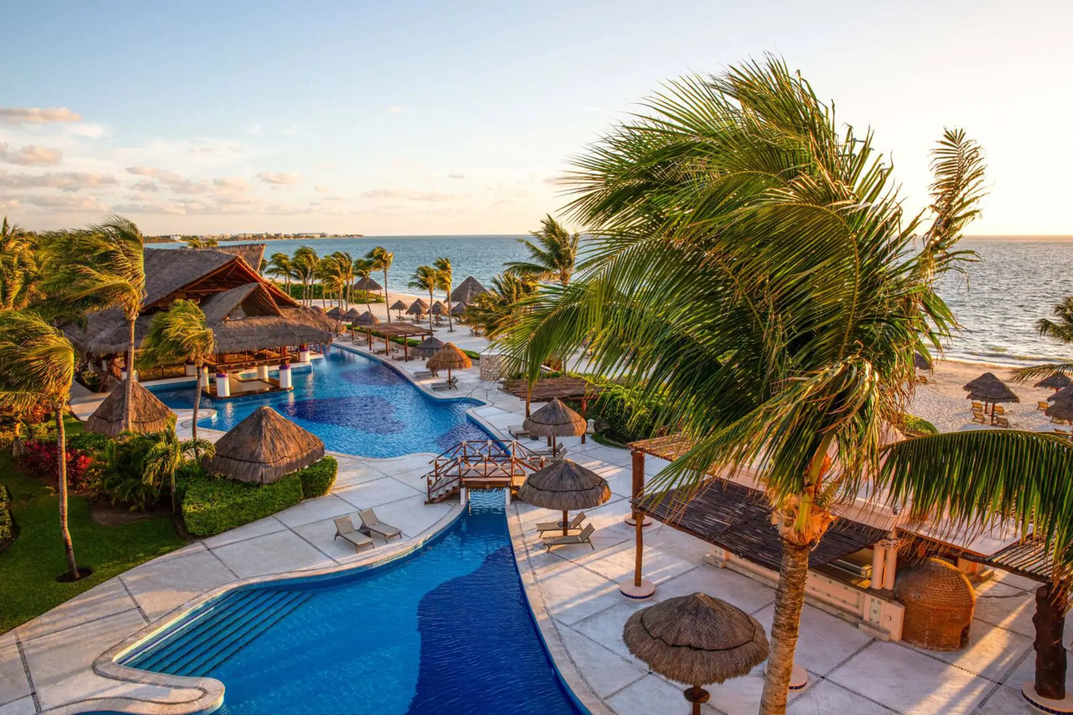 Beachfront infinity pool with palapa umbrellas, wooden bridges, and Caribbean views during golden hour.