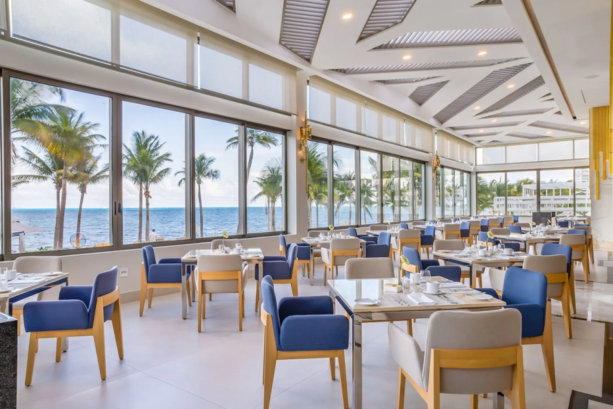 Bright oceanview restaurant with floor-to-ceiling windows, modern furniture, and palm trees in the background.