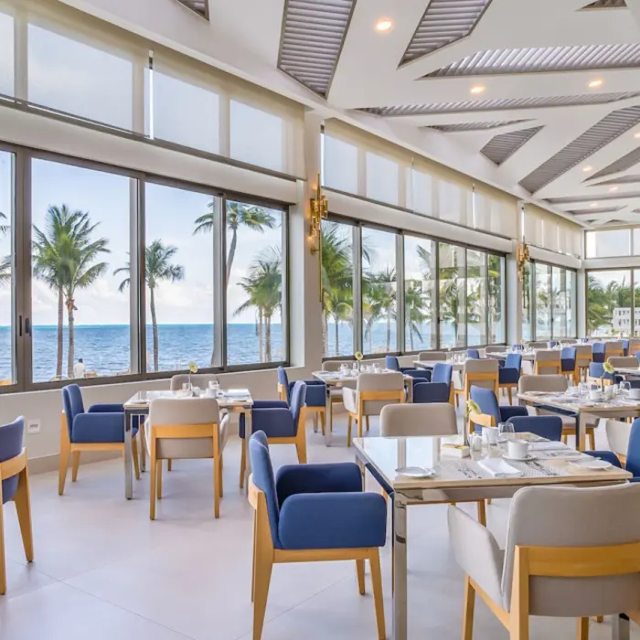 Bright oceanview restaurant with floor-to-ceiling windows, modern furniture, and palm trees in the background.
