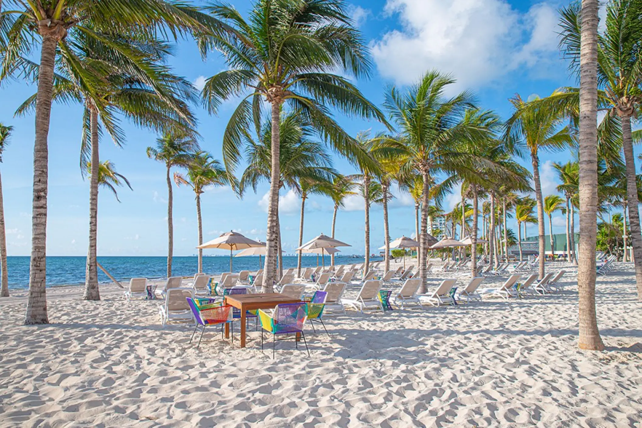 White-sand beach with lounge chairs, umbrellas, palm trees, and ocean views under a clear blue sky.