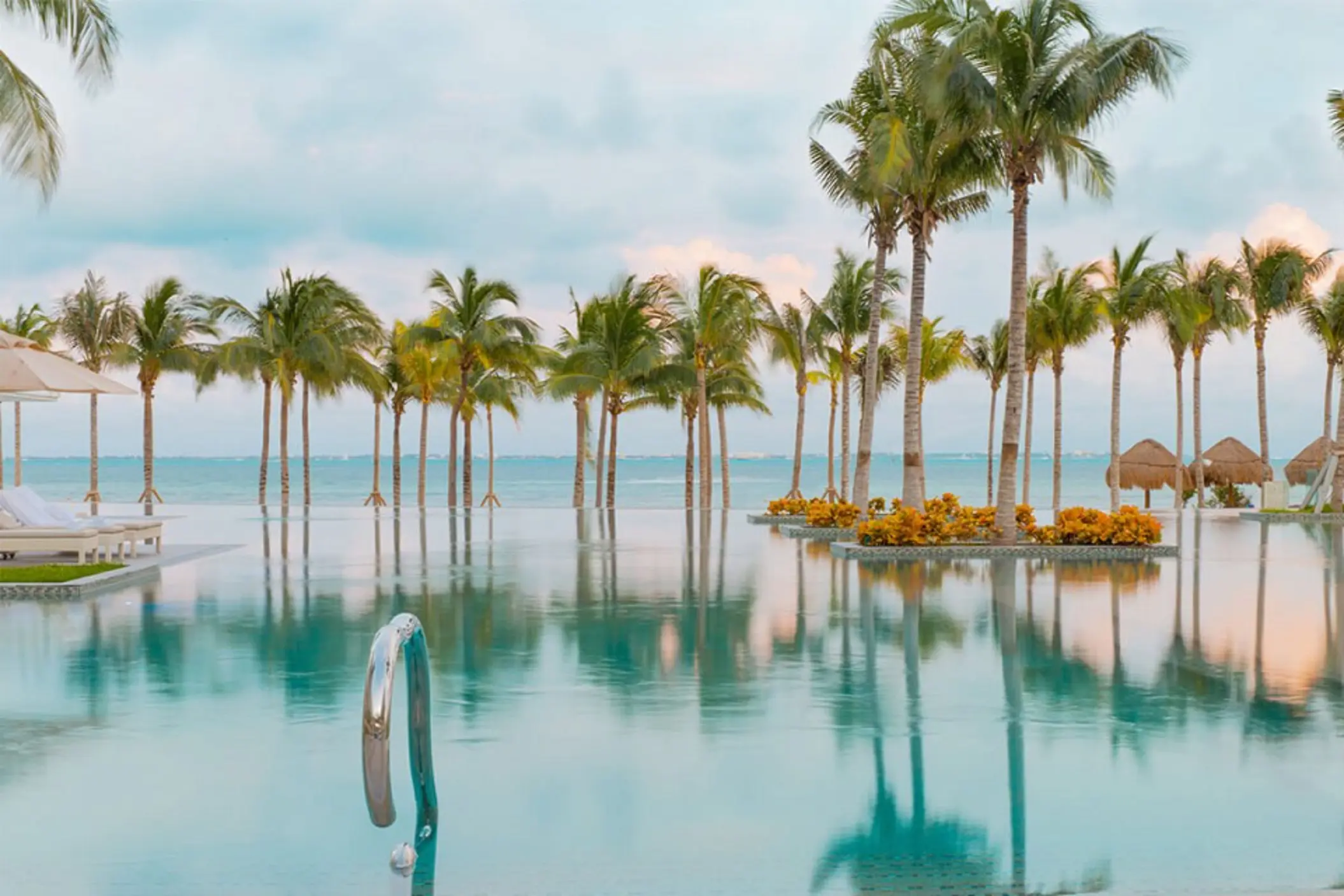 Infinity pool with ocean views, palm trees reflected on the water, and sun loungers under umbrellas.