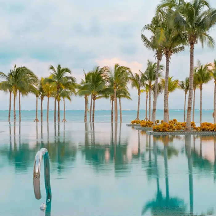 Infinity pool with ocean views, palm trees reflected on the water, and sun loungers under umbrellas.